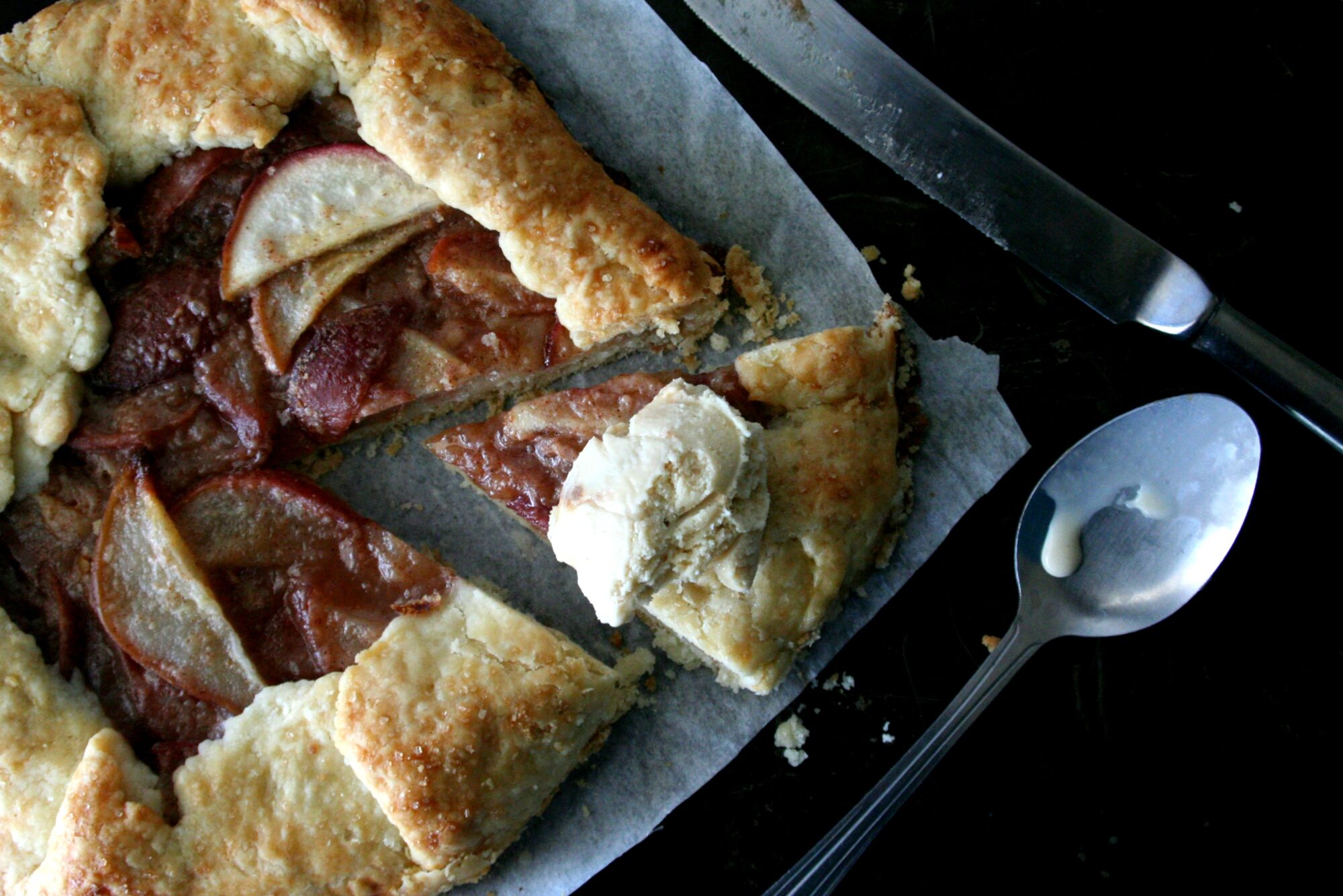 An overhead image of a sliced ginger pear galette with ice cream on a black sheet pan next to a knife and spoon.