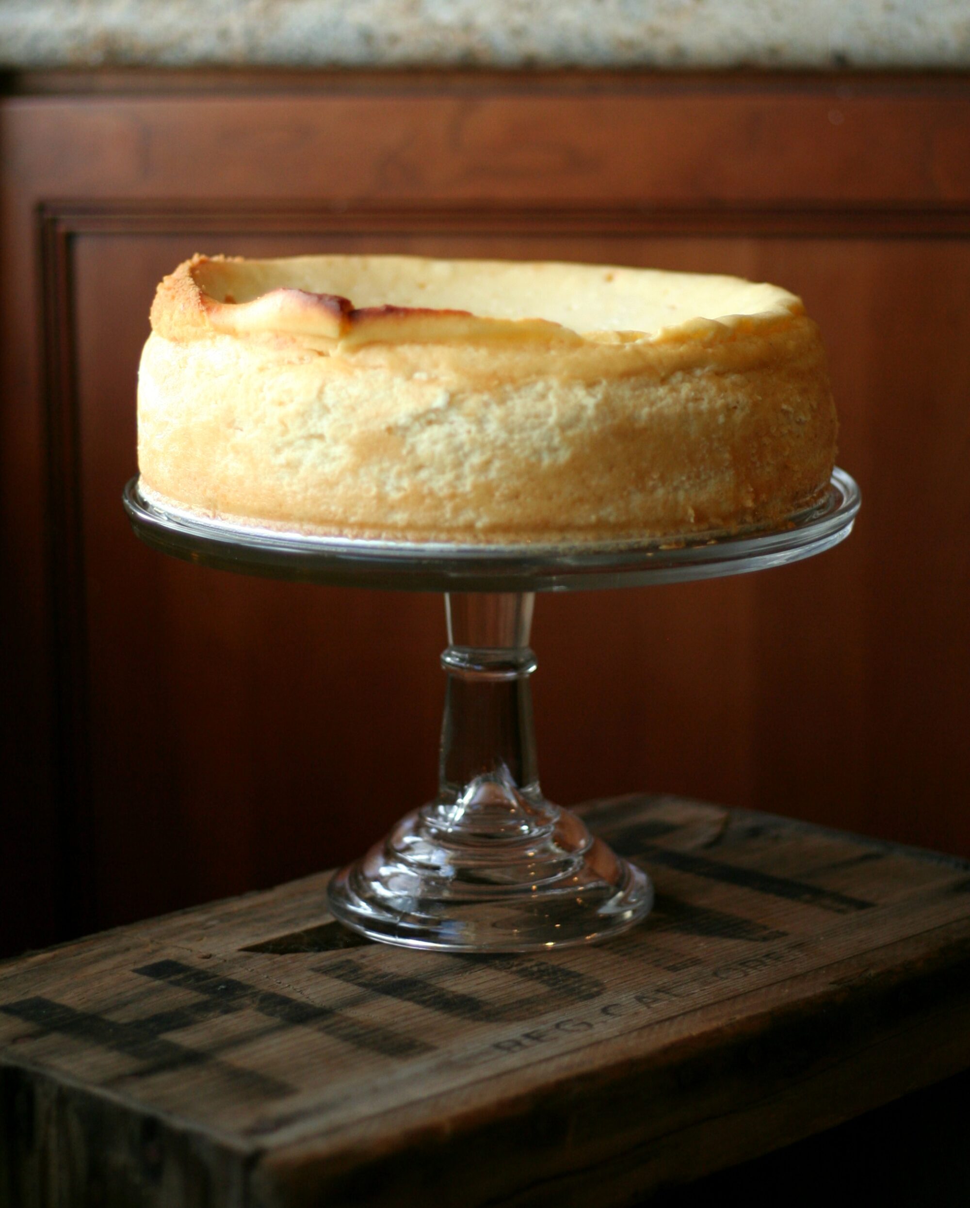 A side image of a mascarpone cheesecake with almond cookie crust on a clear stand with a dark wood background.