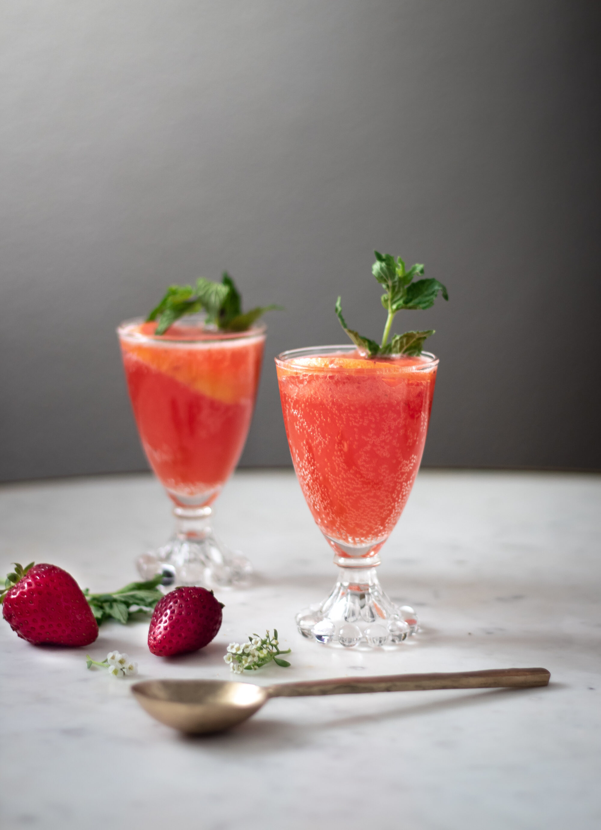 Two glasses of sparkling strawberry bourbon lemonade on a white counter next to strawberries and white blossoms.