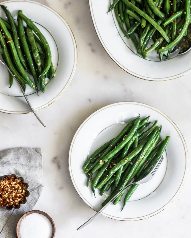 An overhead image of two white plates of green beans on a marble counter next to an oval white tray of green beans, a beige linen, and a bowl of pepper flakes.