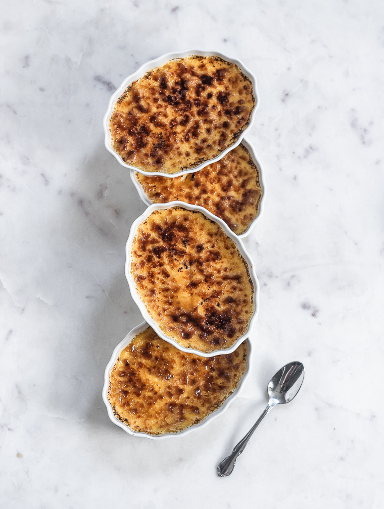 An overhead image of three oval dishes of orange liqueur and cardamom crème brûlée topped with two more creme brulees on a marble counter next to a silver spoon.