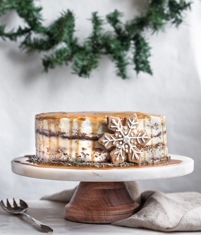 A side image of a naked gingerbread cake drizzled with caramel and decorated with a snowflake gingerbread cookie on a wood cake stand, placed on a marble counter with garland in the background.