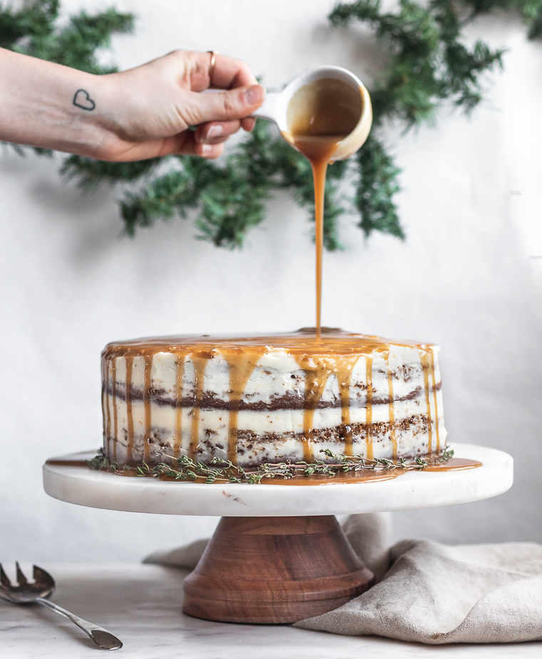 A side image of a woman's hand drizzling caramel over a gingerbread cake on a wood cake stand, placed on a marble counter with garland in the background.