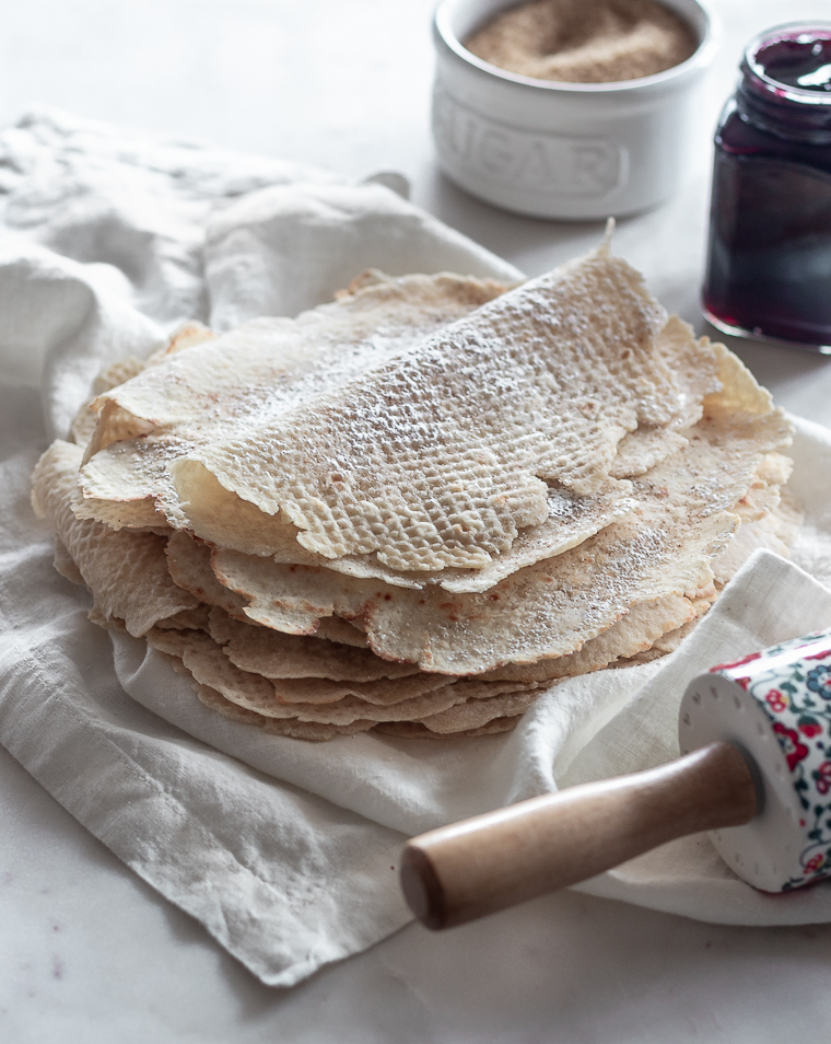 A side image of a stack of lefse on a white linen with a jar of purple jam and a white bowl of Demerara sugar in the background.
