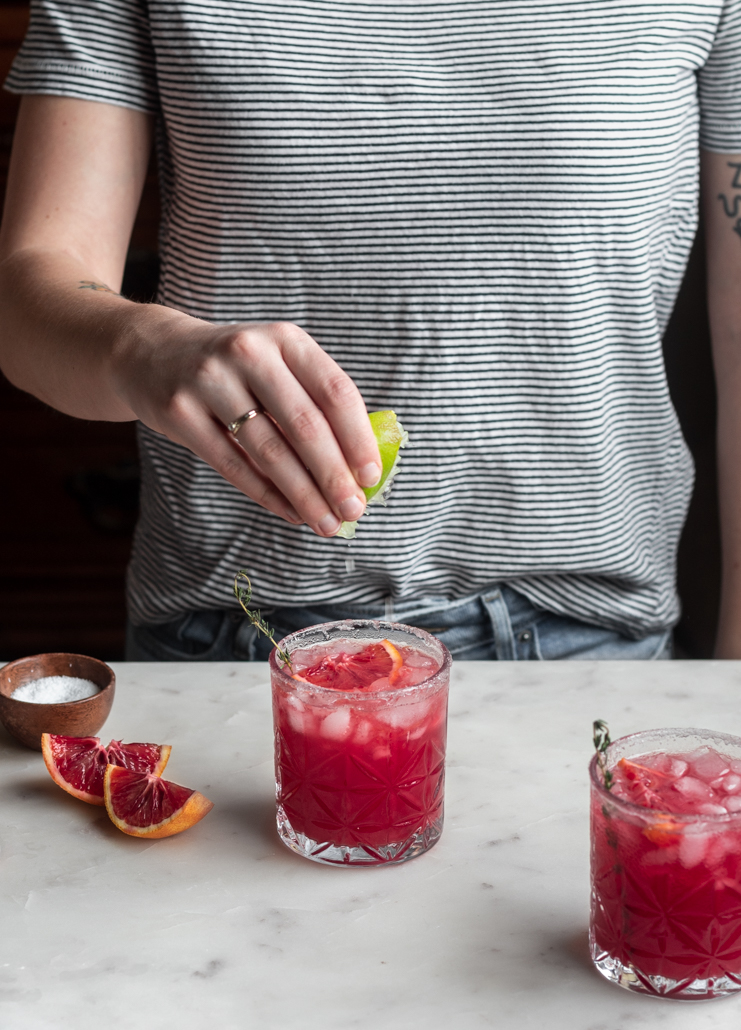 A woman wearing a white and black striped shirt squeezing lime juice into a blood orange margarita on a white marble counter next to a wood bowl of salt and slice of blood orange.