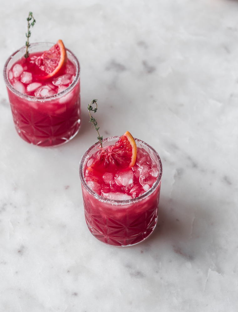 A side image of two blood orange margaritas garnished with slices of orange and sprigs of thyme on a white marble counter.