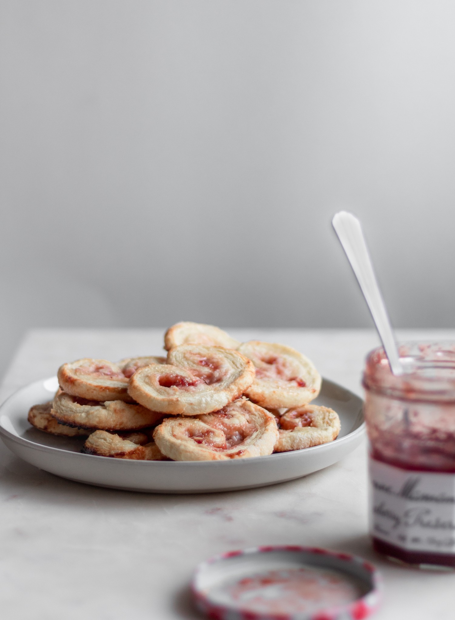 A white plate with a pile of strawberry palmiers with cream cheese filling on a white counter with a jar of jam in the forefront.