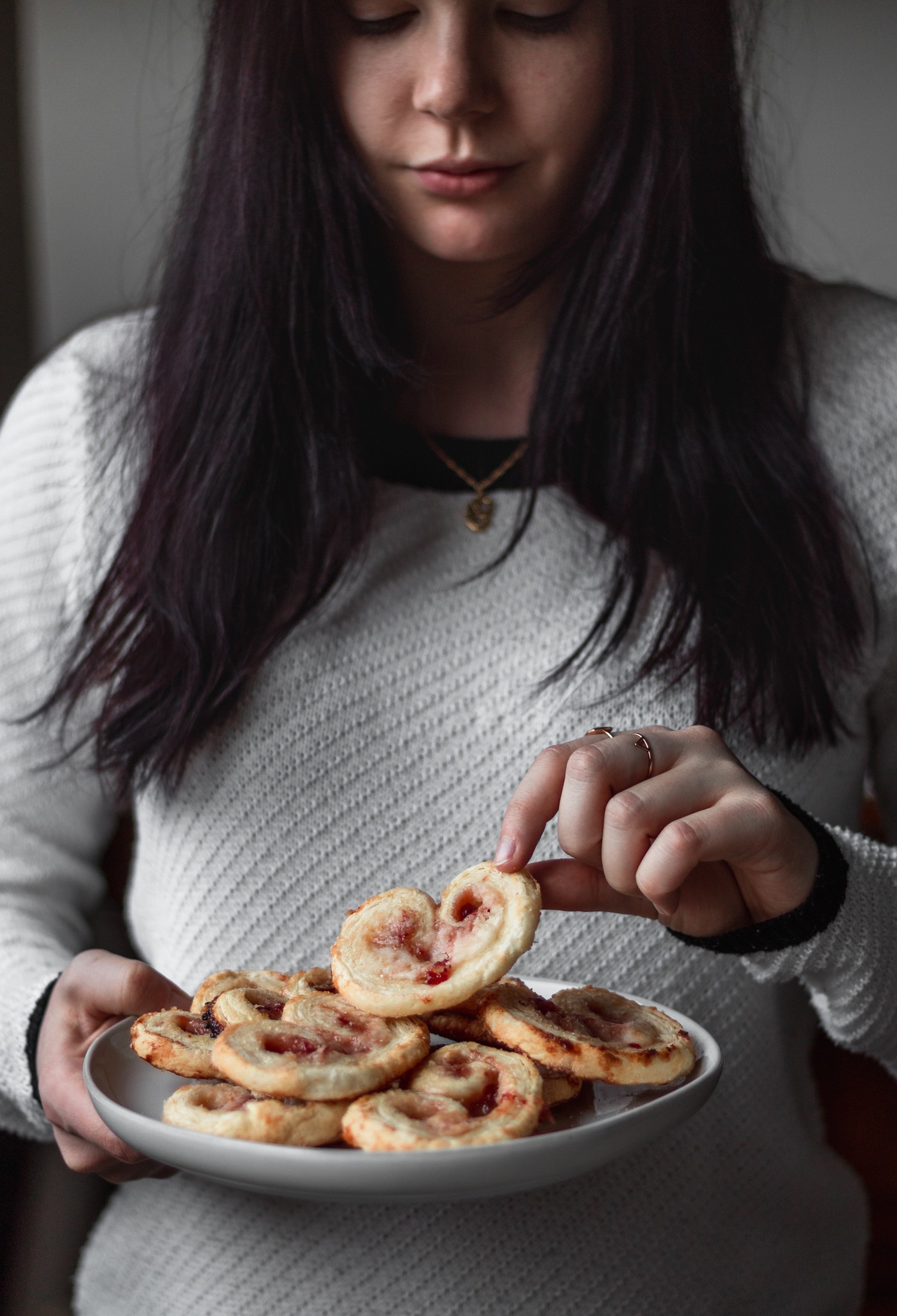 A side image of a woman with dark red hair wearing a white sweater holding a plate of elephant ear cookies.