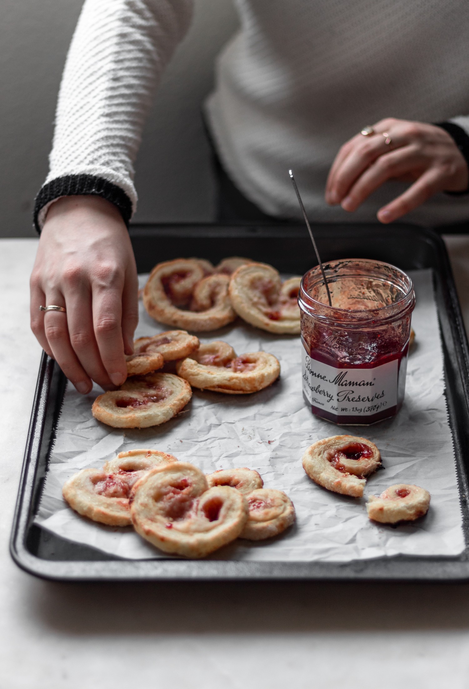 A side image of a woman placing elephant ear cookies on a black sheet pan next to a jar of jam on a white counter.