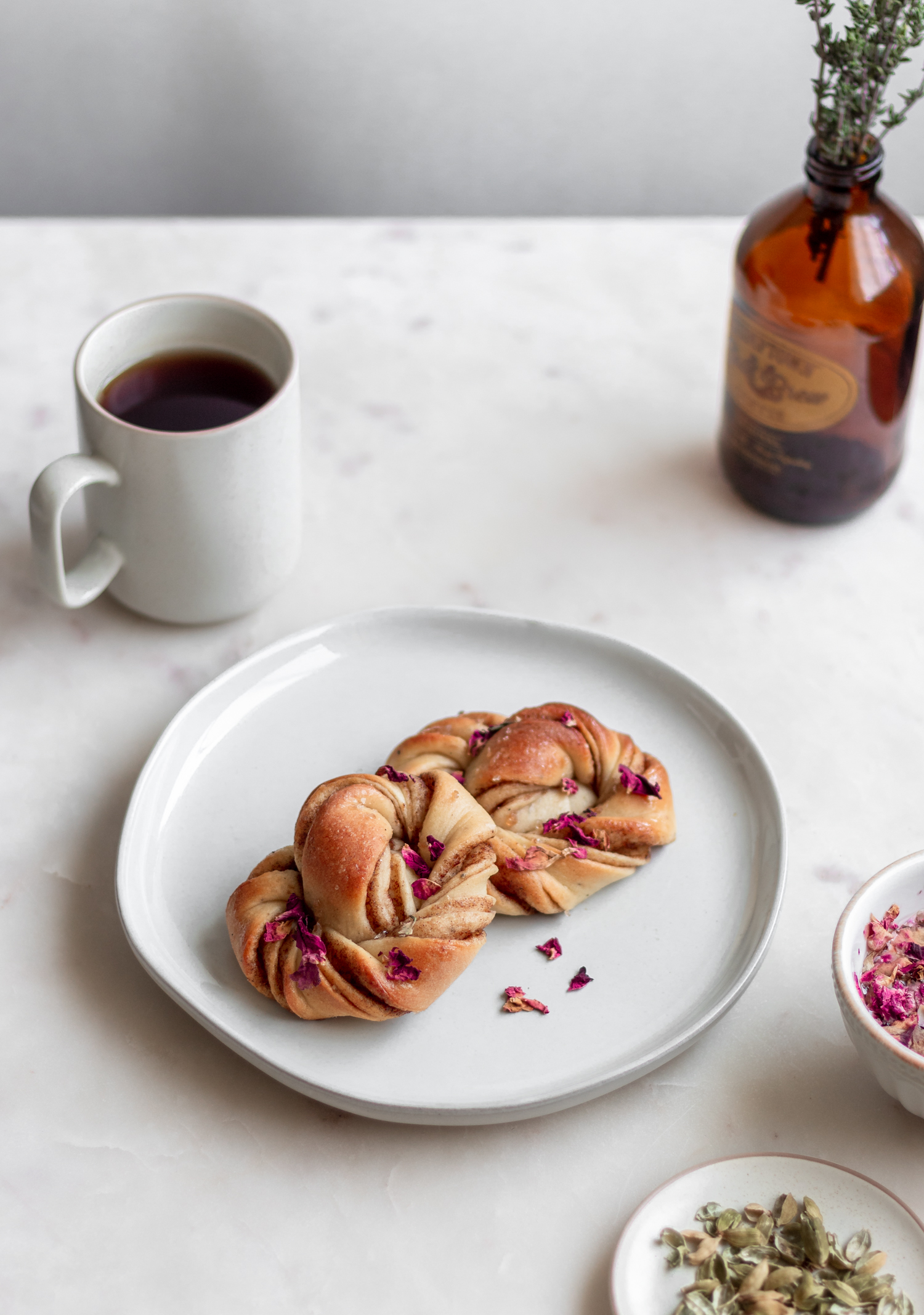 A side image of two Swedish cinnamon rolls with rose petals on a white plate placed on a white counter next to a cup of coffee, white bowl of rose petals, and white bowl of cardamom pods.