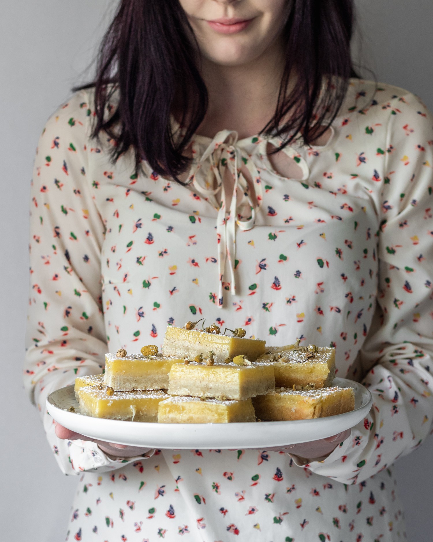 A side image of a woman wearing a white floral shirt holding a white plate of chamomile lemon bars.