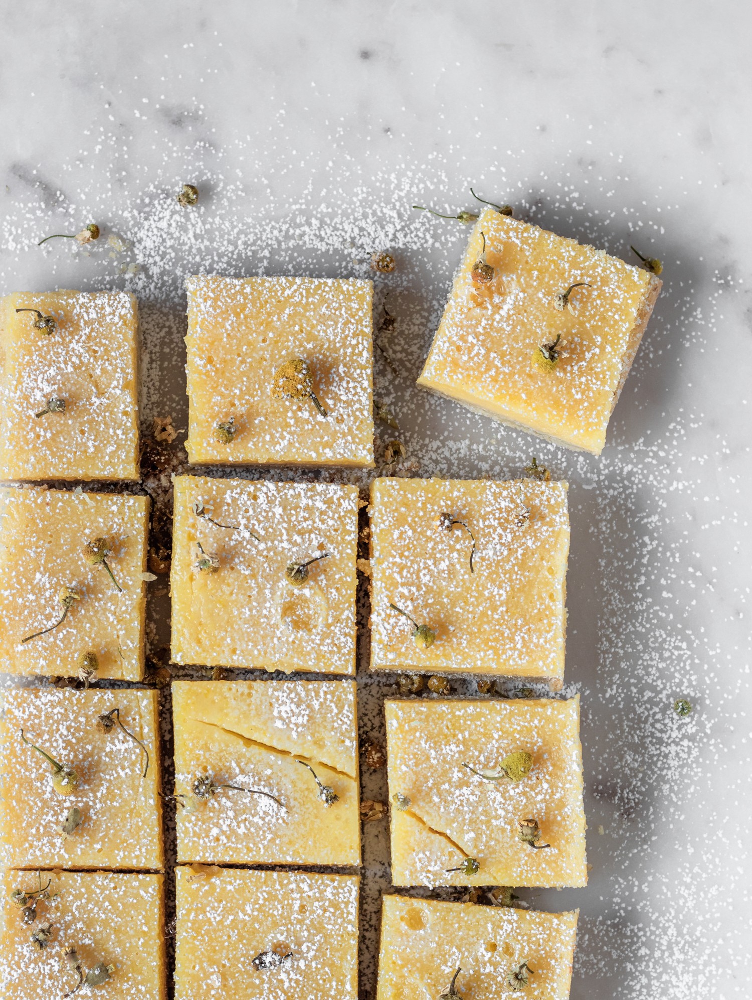 An overhead photo of rows of chamomile lemon bars dusted with powdered sugar on a marble table.