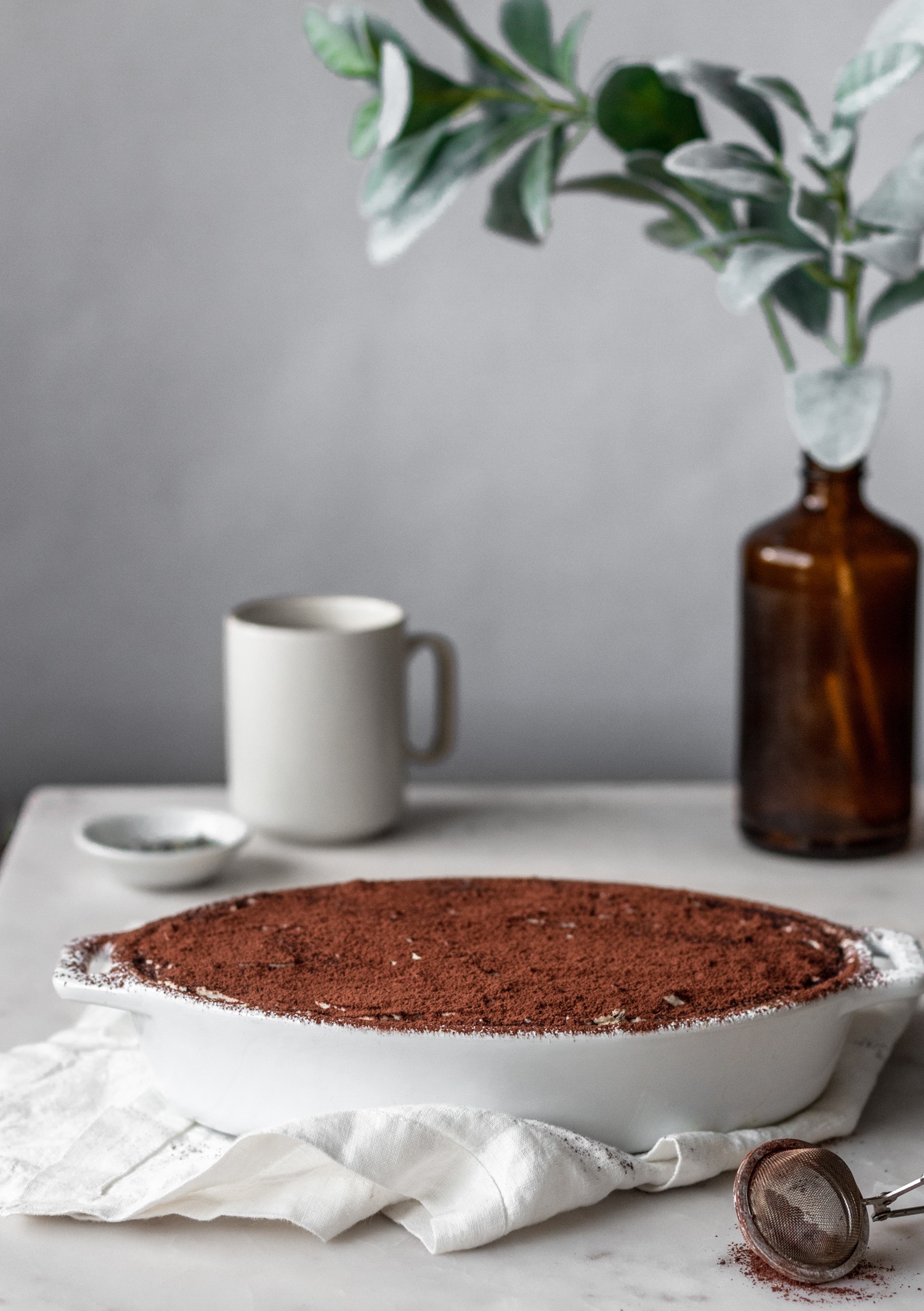 A white dish of London fog tiramisu on a white counter with eucalyptus leaves and a cup of tea in the background.