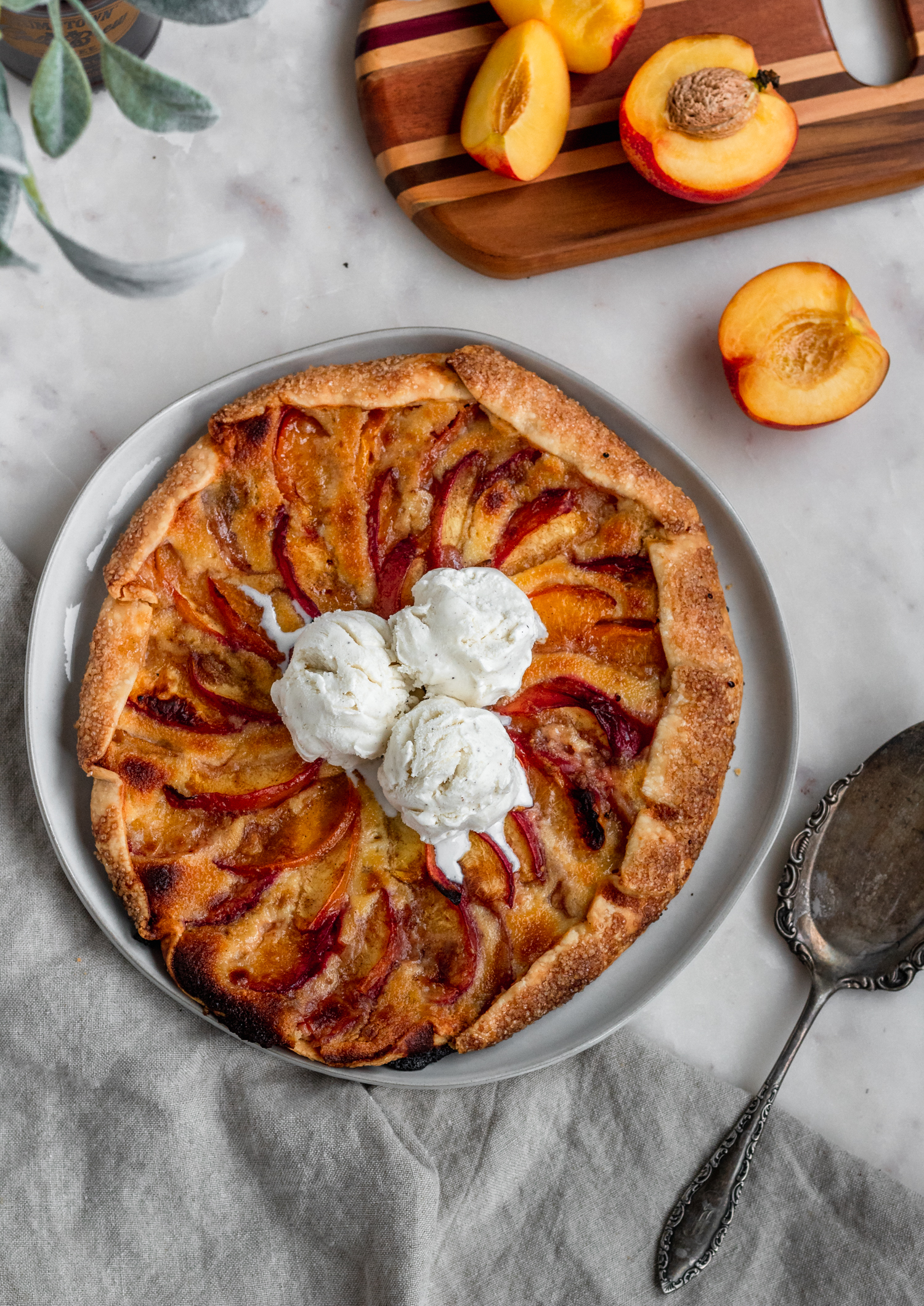 An overhead image of a white plate with a nectarine galette on a white counter next to a wood cutting board with halved peaches and eucalyptus leaves.