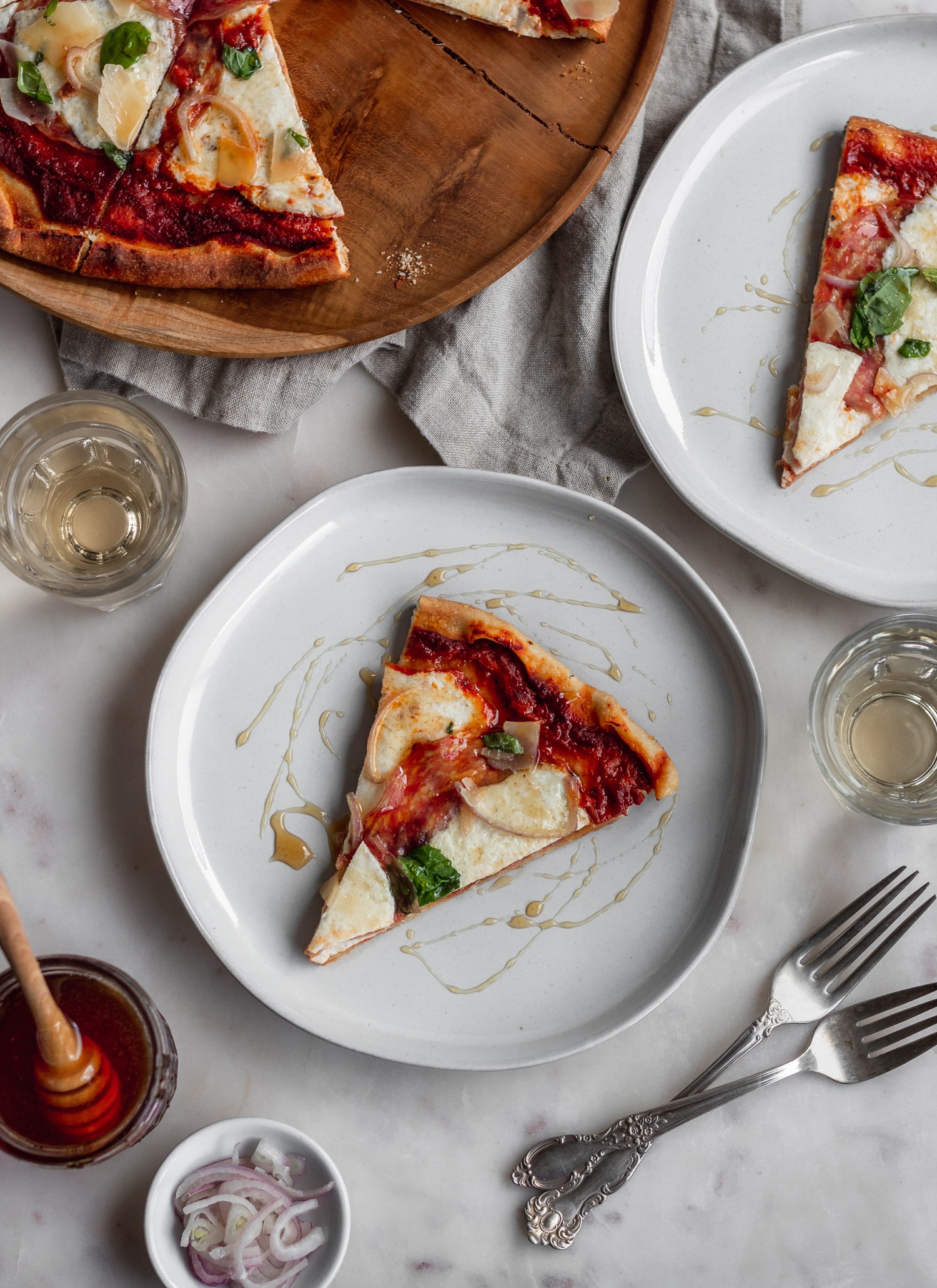 A slice of pizza with soppressata on a white plate next to another plate of pizza, wine glasses, and forks with a marble background.