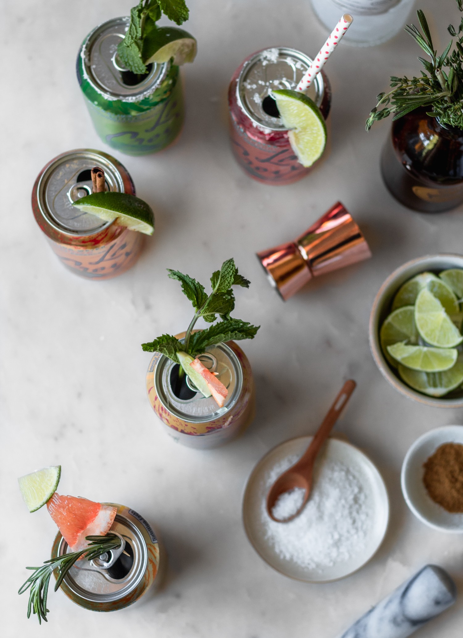 An overhead image of five cans of La Croix with garnishes on a marble counter next to a white bowl of limes, jar of herbs, and bottles of alcohol.