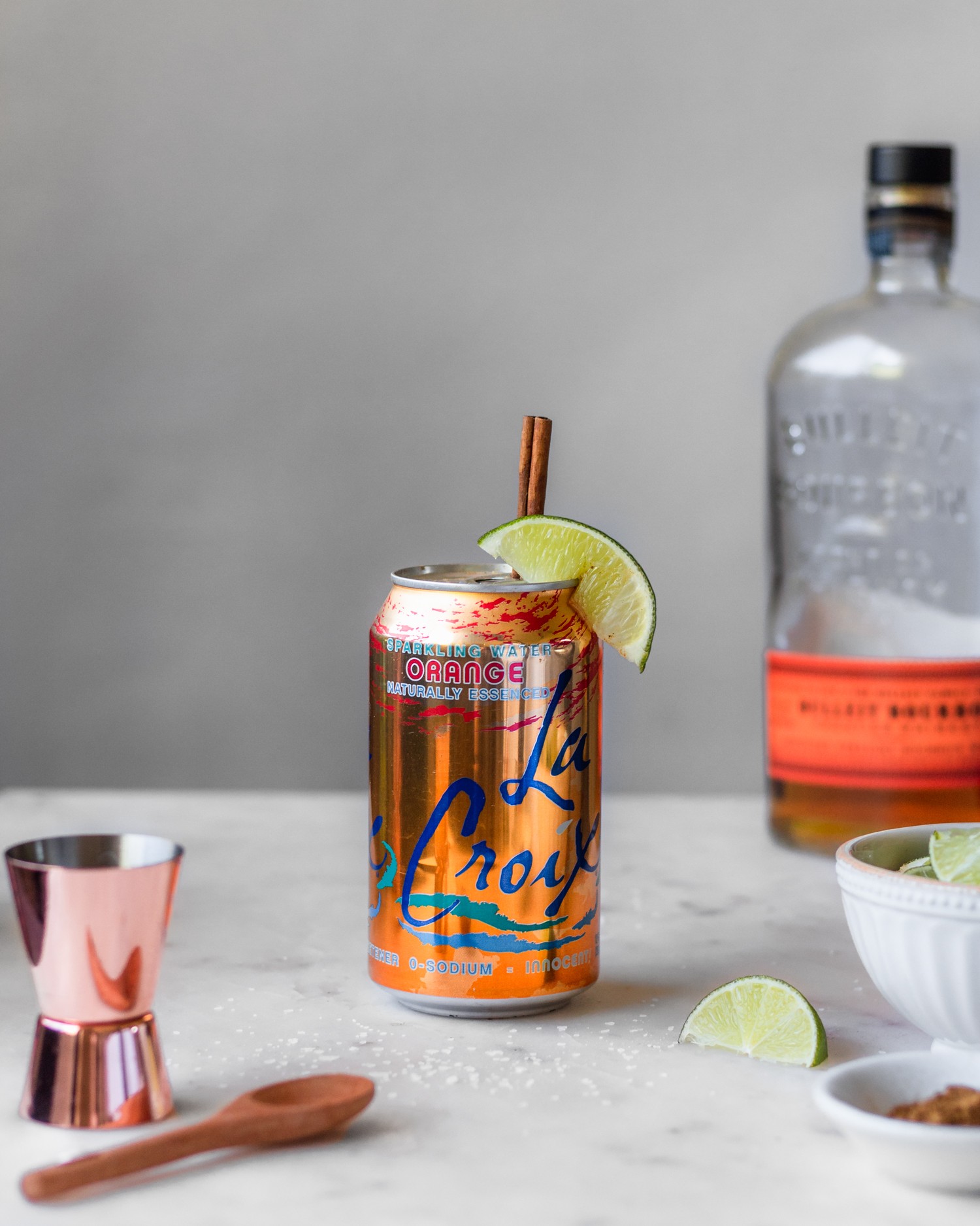 A side image of a can of orange sparkling water with a lime wedge and cinnamon stick next to a bottle of bourbon, white bowl of limes, copper shot glass, and cinnamon on a white counter.