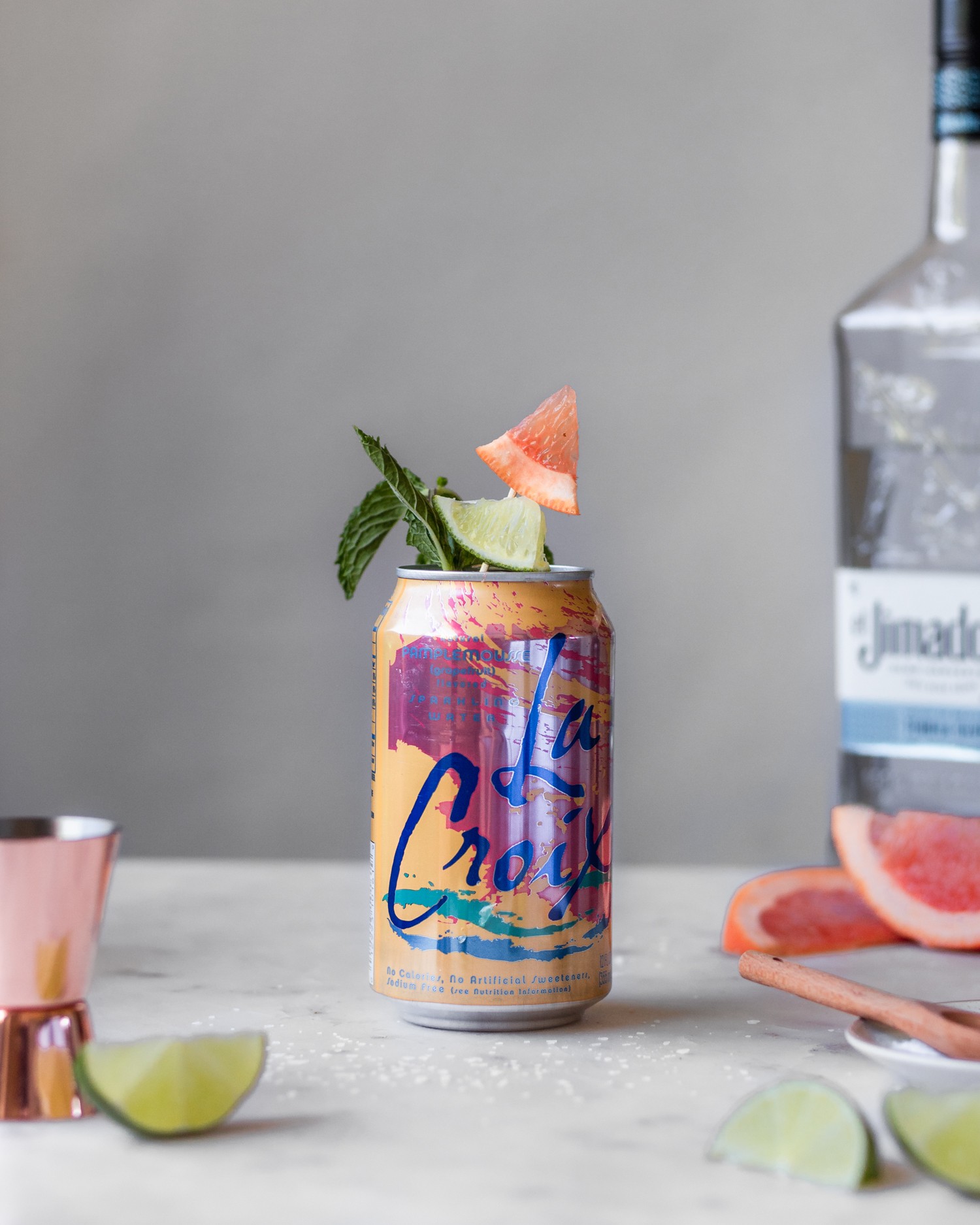 A can of grapefruit sparkling water garnished with citrus and mint on a white table next to a bottle of tequila, citrus wedges, and copper shot glass.