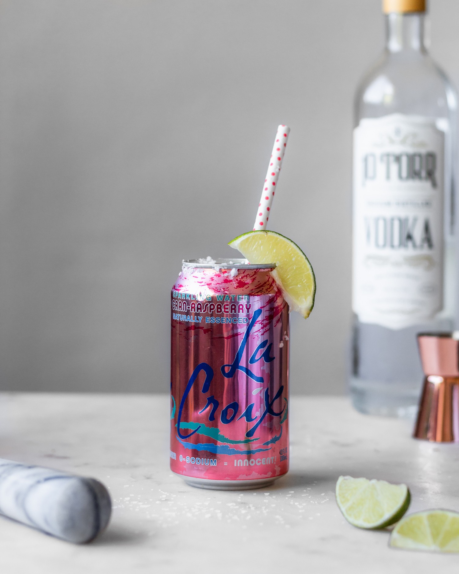 A straight on image of a can of cran-raspberry La Croix with a lime wedge and polka dot straw next to a bottle of vodka, lime wedges, and marble pestle with a white background.