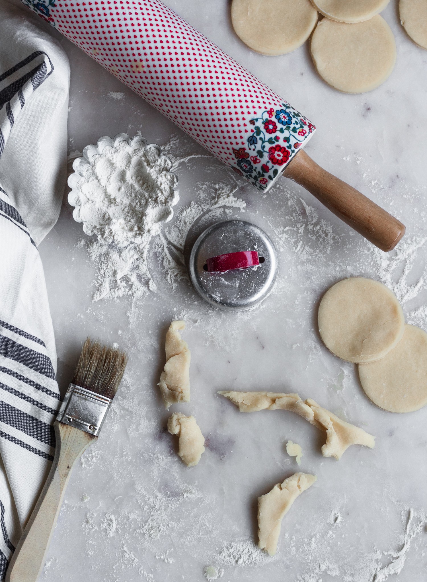 Pie dough rounds on a floured marble counter next to a white linen and flowery rolling pin.