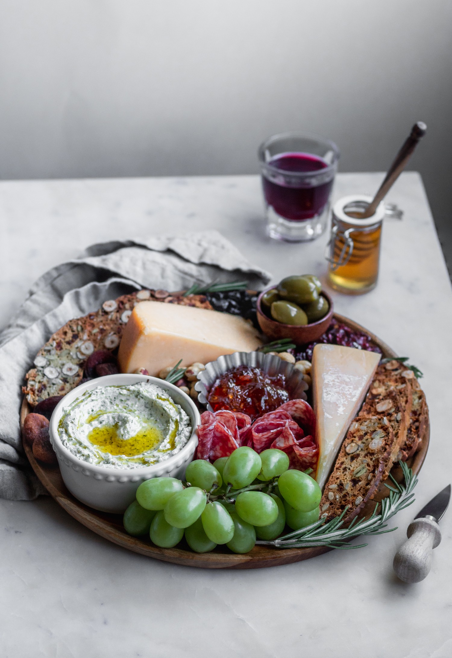 A side image of a cheese board on a wood circular plate placed on a marble counter next to a glass of red wine and jar of honey.