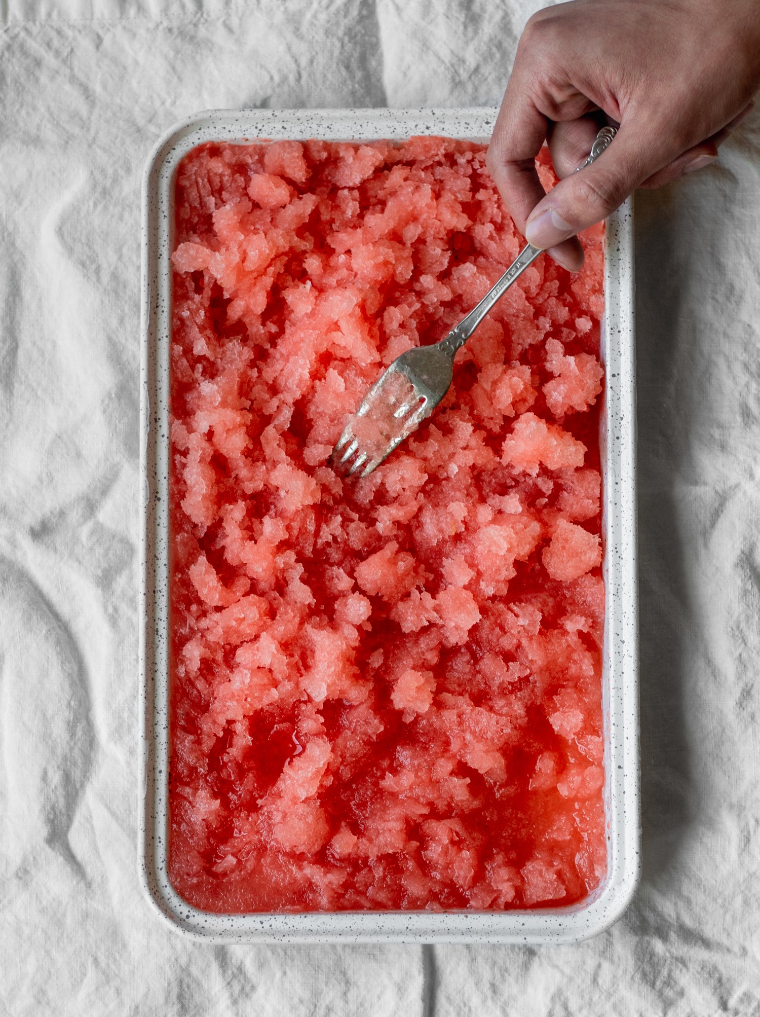 An overhead image of a strawberry aperol spritz granita in a white tin on a white table cloth with a man's hand scraping the granita.