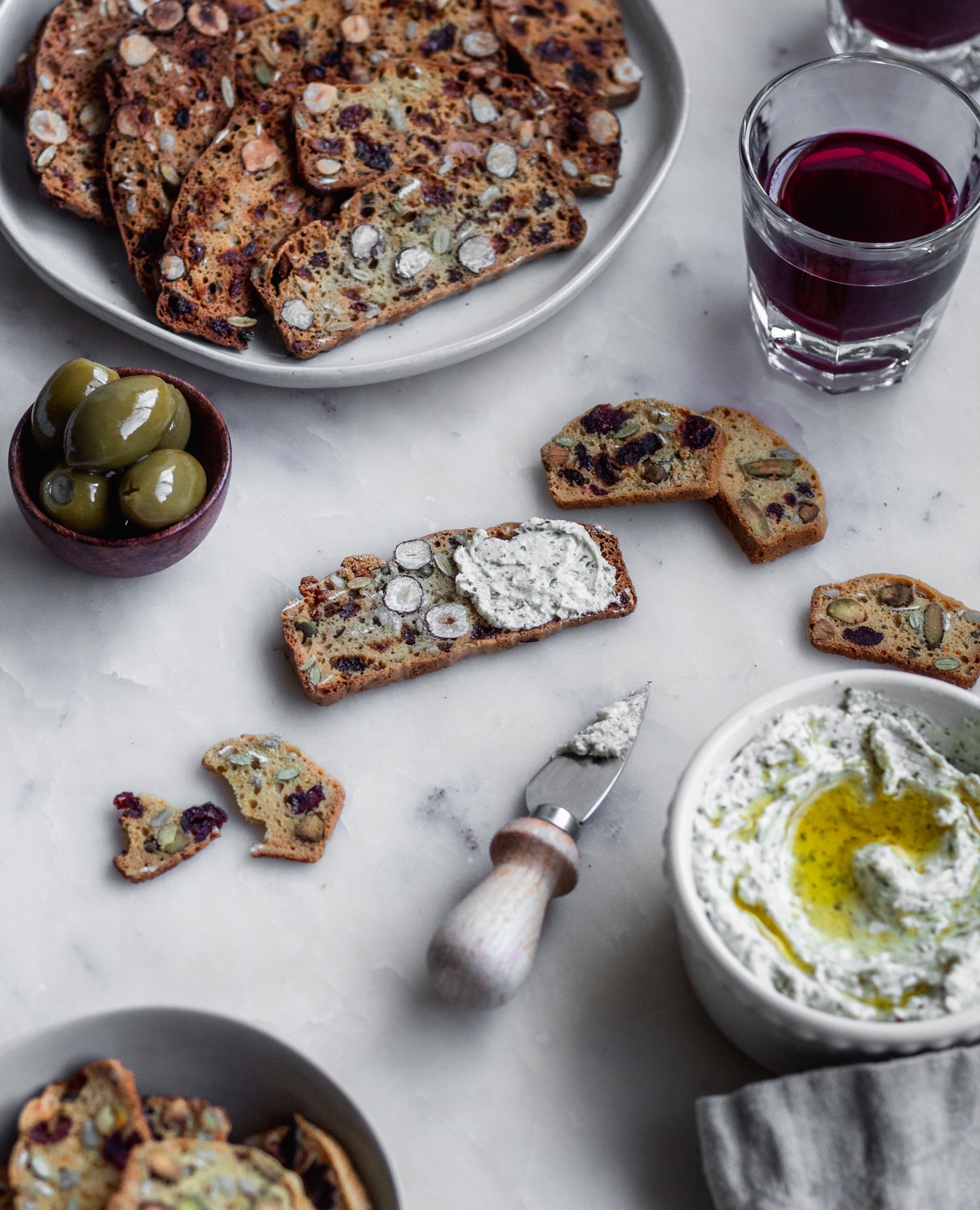 A side image of a homemade fruit and nut crisp on a white marble counter surrounded by more crackers, a bowl of whipped cheese, a white plate of crackers, a glass of red wine, and a wood bowl of olives.