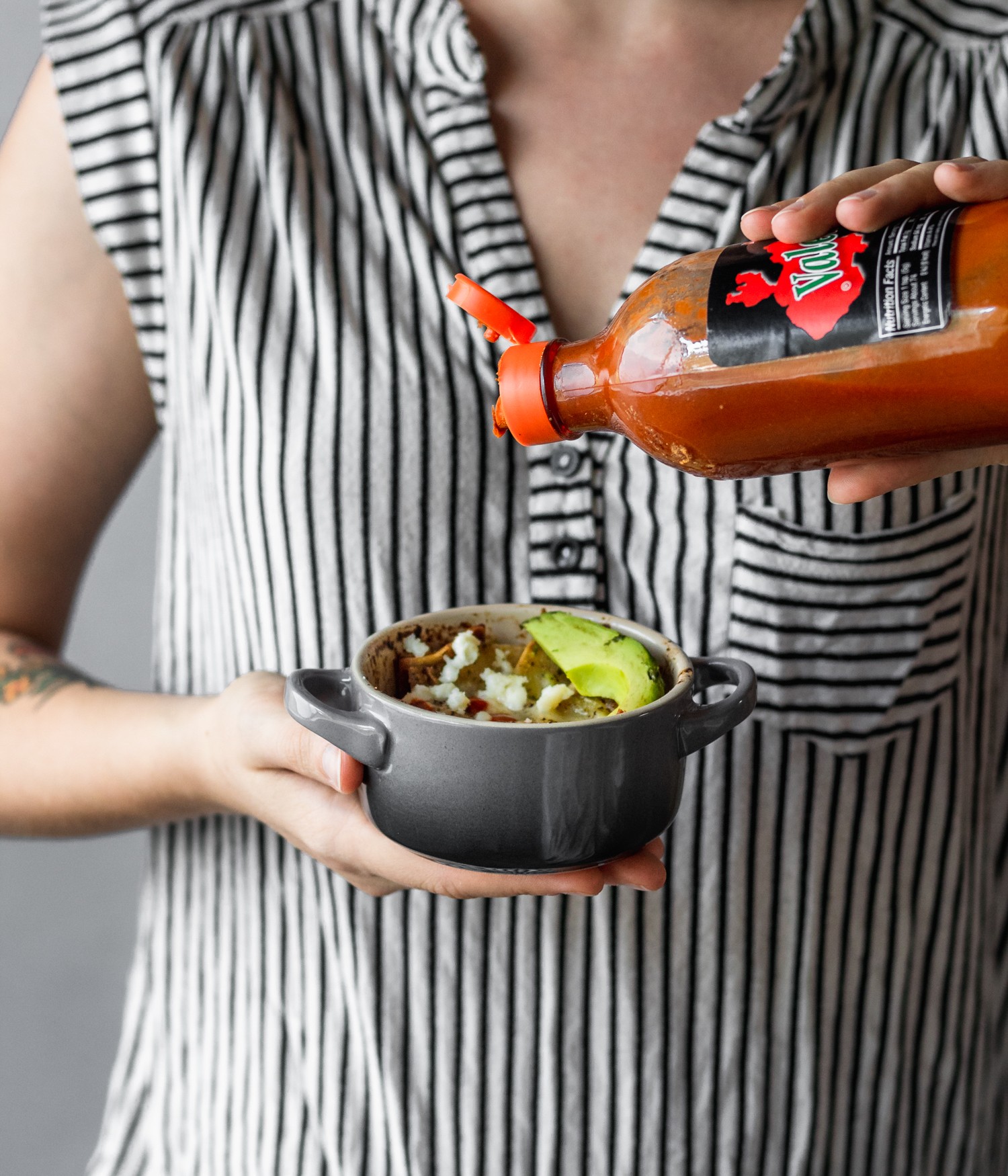 A woman wearing a black and white striped shirt pouring Valentina over a ramekin with eggs, avocado, and green salsa.