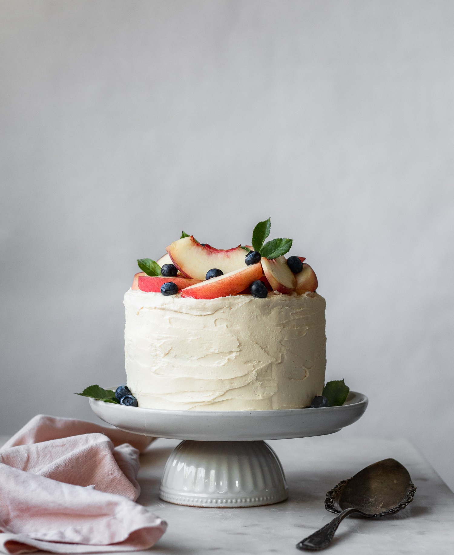 A vanilla cake with peach filling and caramelized white chocolate buttercream decorated with peaches and blueberries on a white cake stand with a white background.