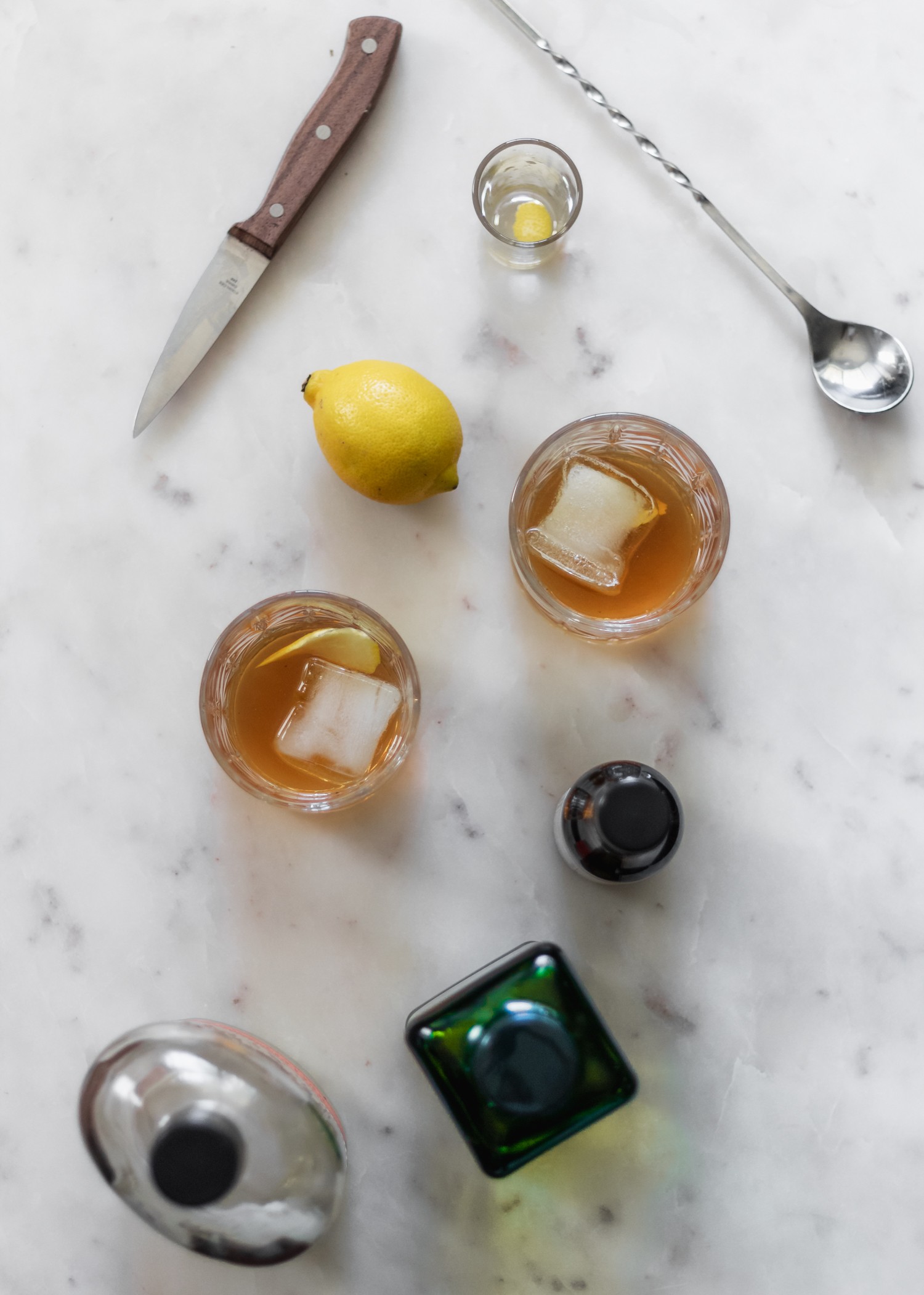 An overhead image of two ginger old fashioneds on a marble counter next to a lemon, bar spoon, green bottle, and bottle of bitters.