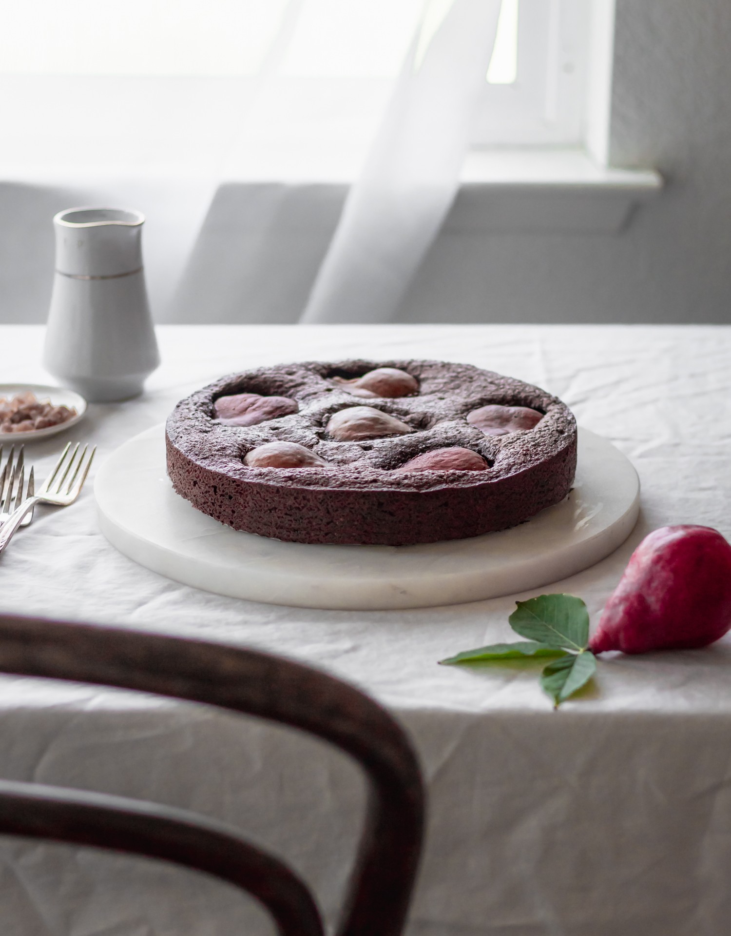 A side image of a flourless chocolate cake with red pears on a marble cake plate on a linen next to a red pear and vintage forks. In the background is a window with a white curtain
