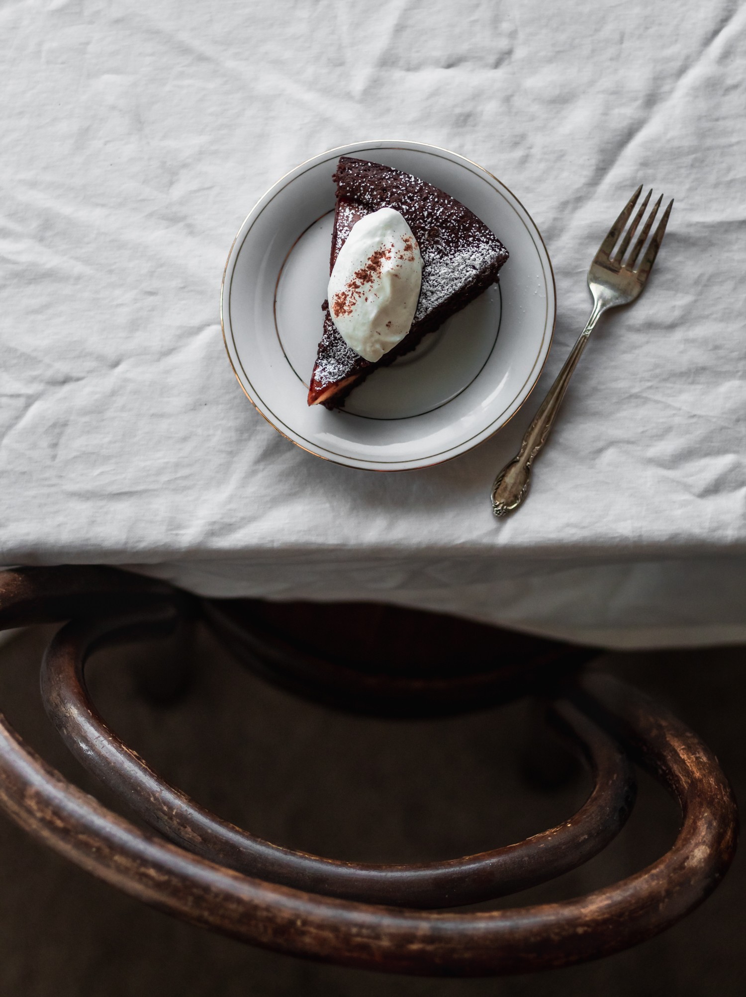 An overhead image of a white plate with a slice of chocolate hazelnut pear cake with whipped cream on a white linen next to a vintage fork. In the bottom of the photo is a vintage wooden chair.
