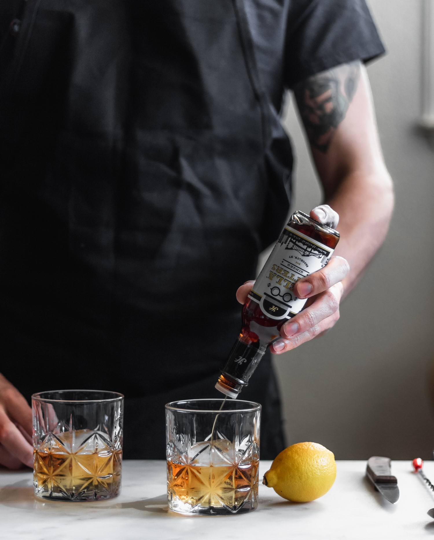 A side image of a man in a black smock pouring ginger bitters over a ginger old fashioned on a marble counter next to a lemon and wood knife.