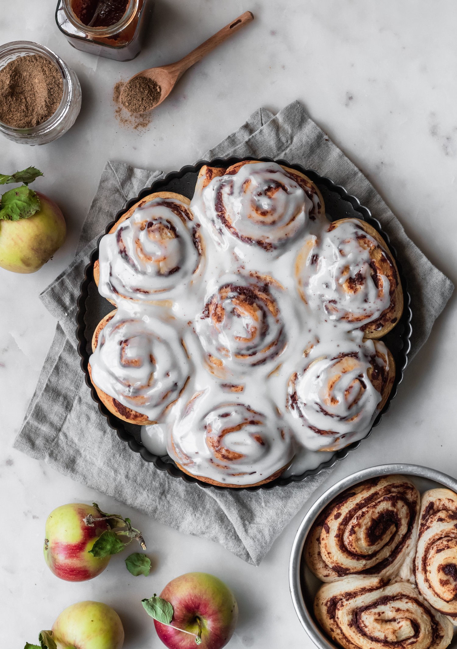 An overhead image of a metal circle pan of apple butter cinnamon rolls with brown butter icing on a marble counter next to a smaller metal tin of un-iced cinnamon rolls, a beige linen, apples, and a jar of apple butter.