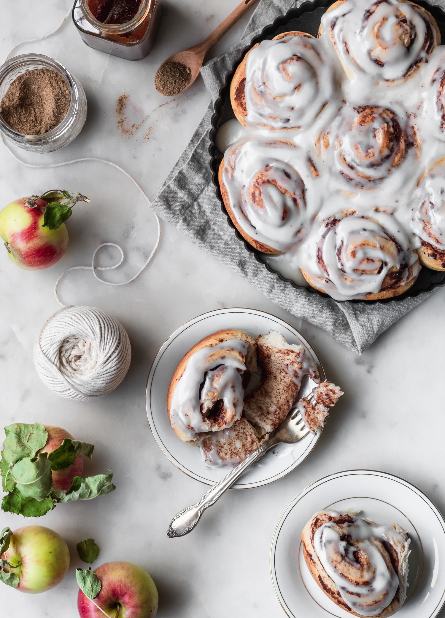 An overhead image of an apple butter cinnamon roll on a white plate with a fork tearing it apart. The plate is on a marble counter next to a larger tray of apple butter cinnamon rolls, apples, and twine.