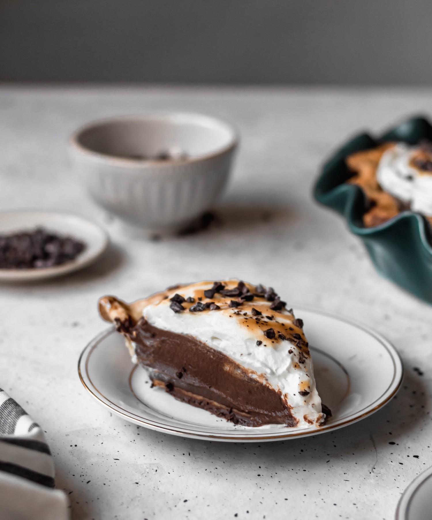 A closeup side image of a slice of chocolate cream pie on a white plate with a white bowl, teal pie plate, and striped linen in the background.