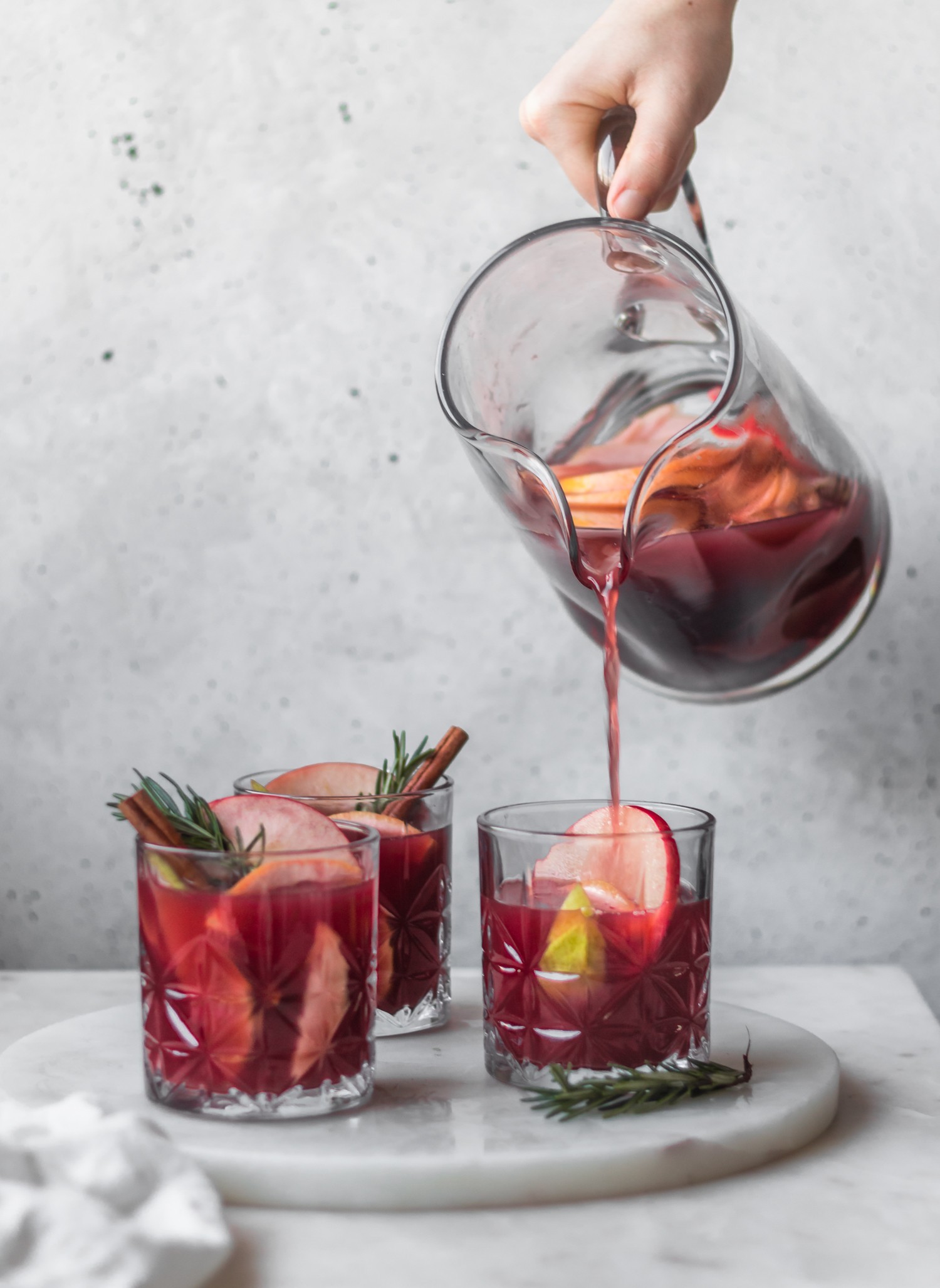 A side image of a woman's hand pouring red wine sangria into a glass of fruit on a marble counter next to two more glasses of sangria.