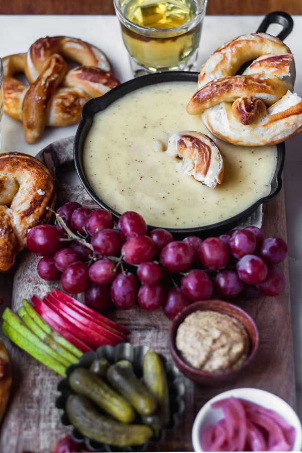 White cheddar and German lager cheese dip in a cast iron skillet with a pretzel being dipped into the center. Fruits, vegetables, and beer surround the skillet.