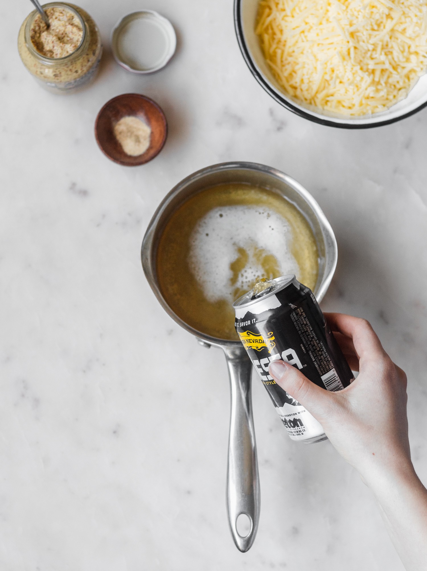 A woman's hand pouring a can of beer into a saucepan placed on a white background. Cheddar cheese, mustard, and spices are surrounding the pan.