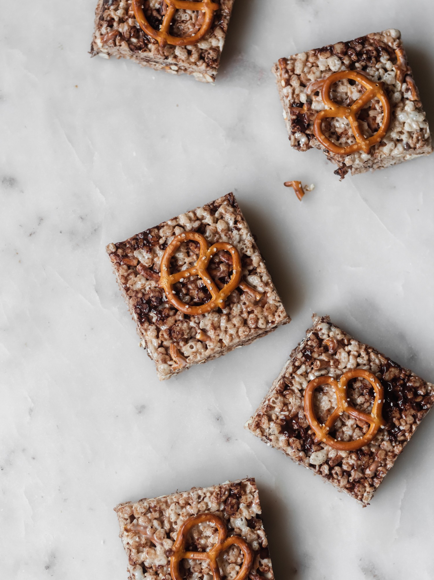 Five cereal bars with chocolate and pretzels on a white marble counter.