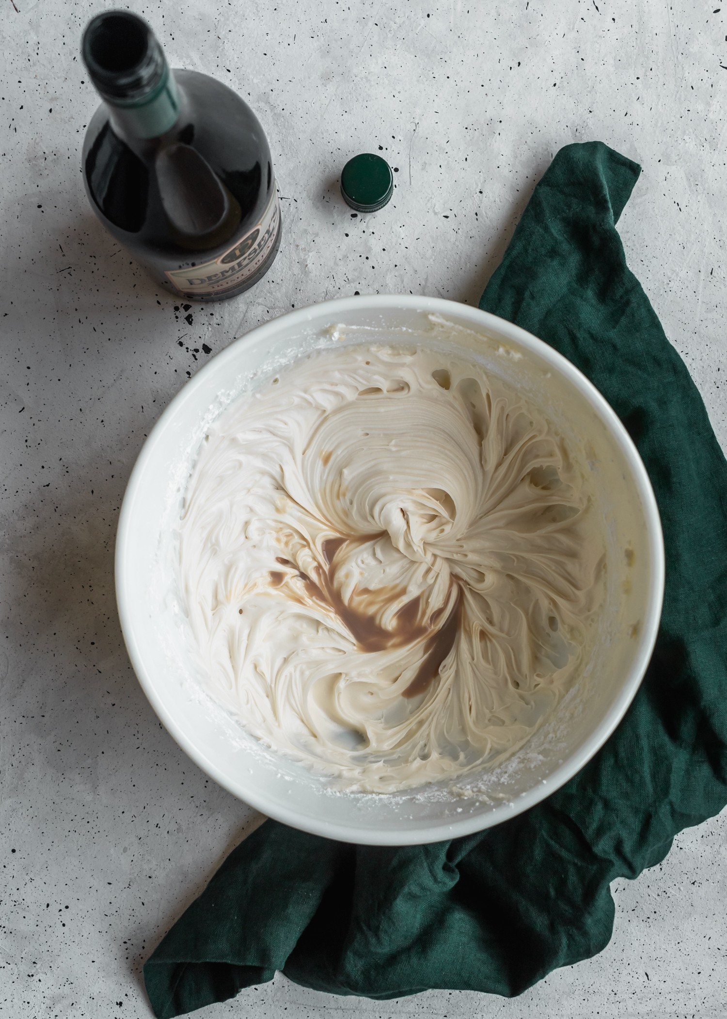 A bowl of cream cheese frosting with a drizzle of Irish cream in the center. There is an opened bottle of Irish cream in the left corner.