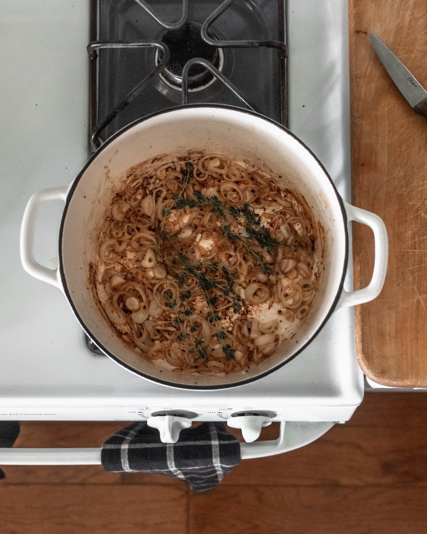 A pot of onions and thyme on a white stove next to a wood counter top.