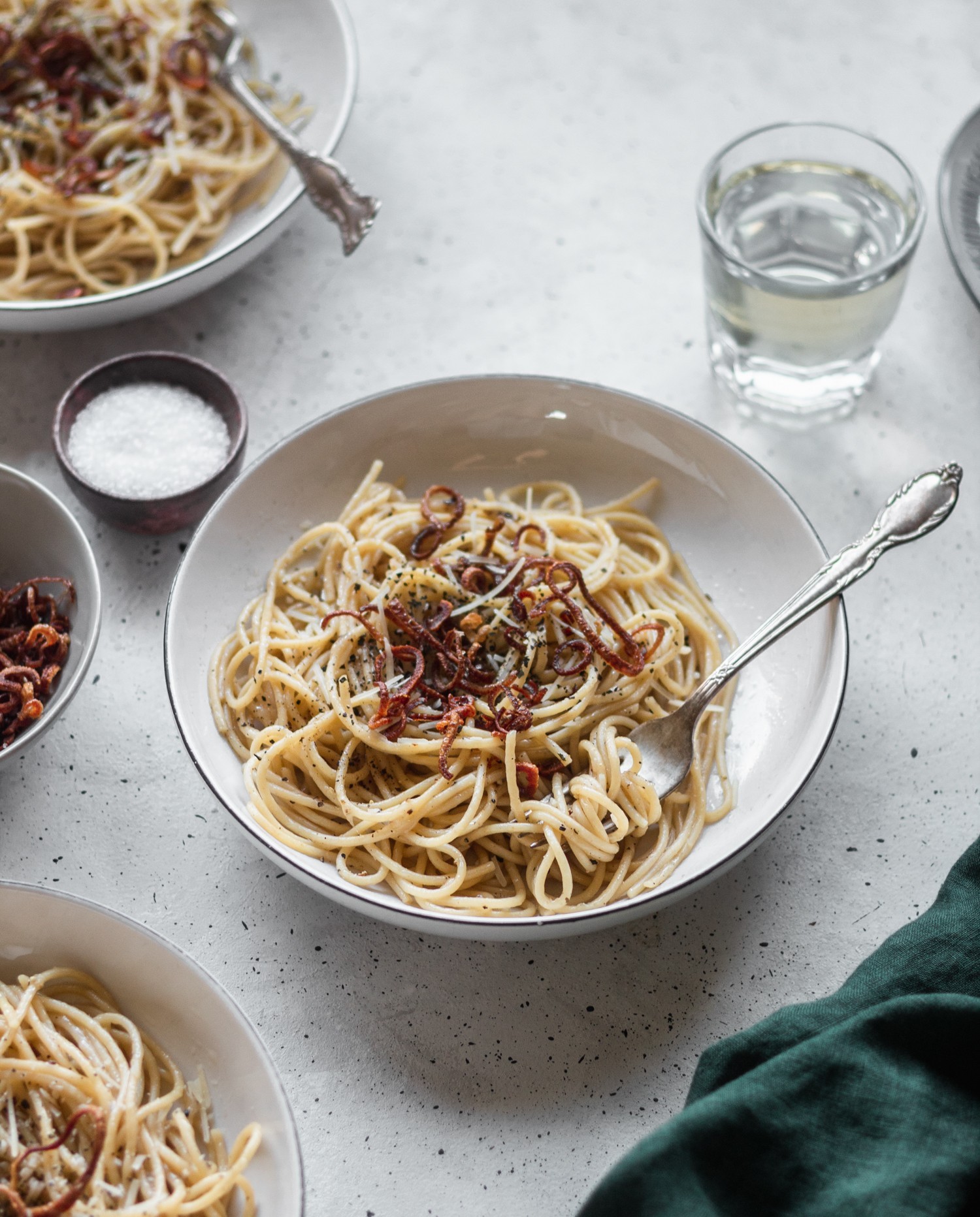 A side shot of a bowl of spaghetti with fried shallots on top, surrounded by a green linen, more bowls of pasta, fried shallots, and a glass of wine on a grey table.