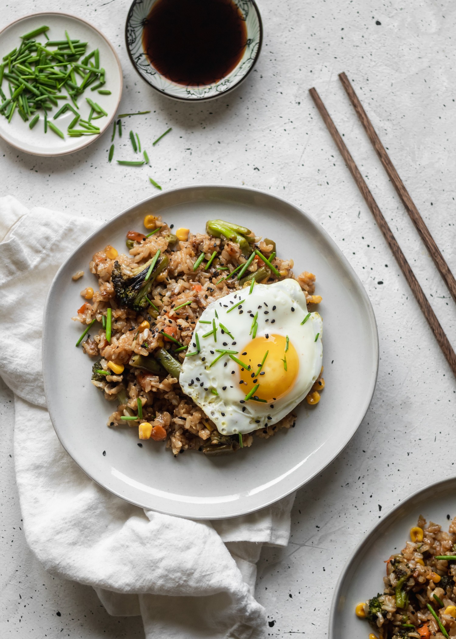 A closeup overhead shot of a plate of fried rice with eggs surrounded by a white linen, chopsticks, a bowl of soy sauce, and a plate of chives on a grey table.