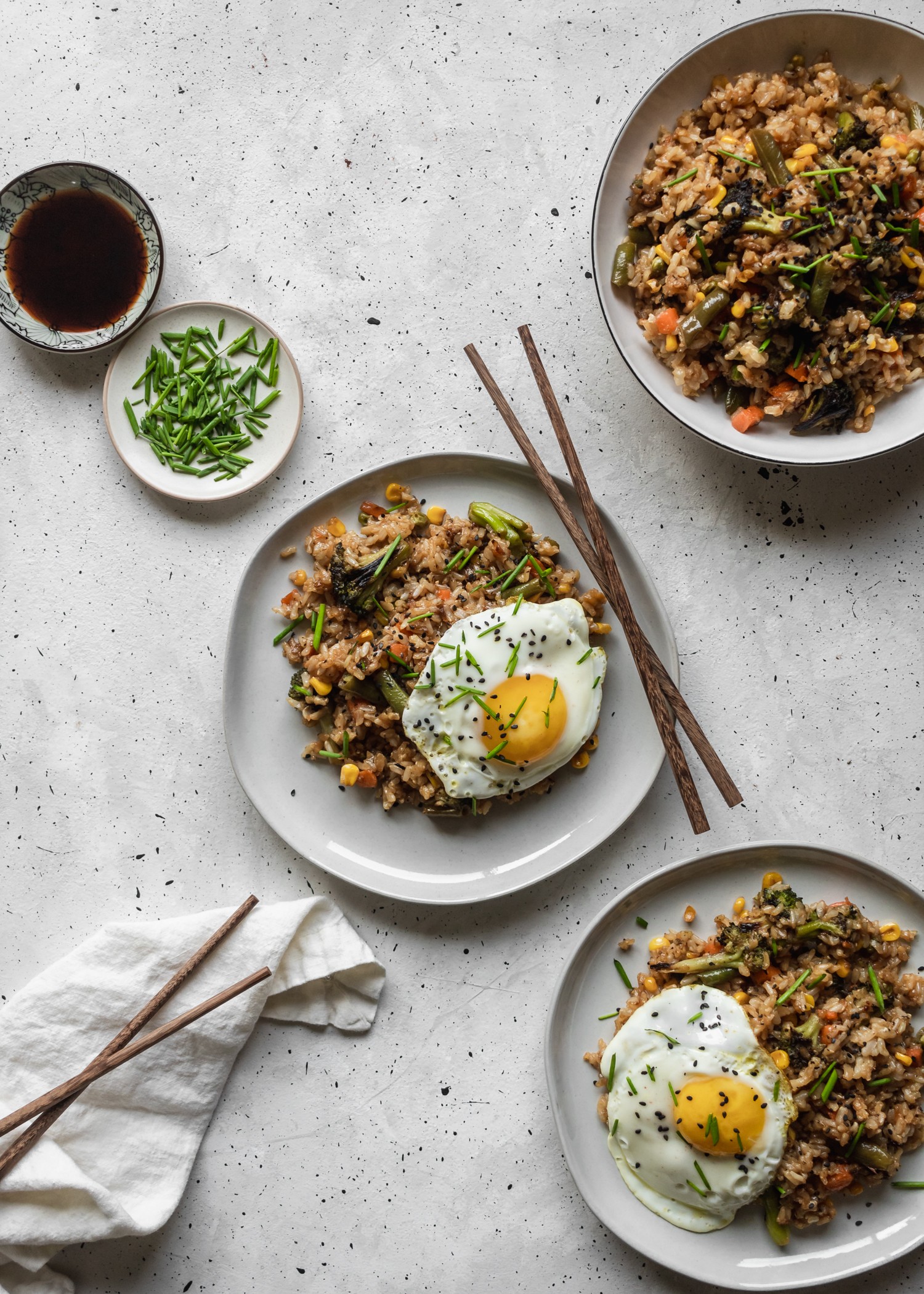 A bird's eye shot of three plates of fried rice next to a white linen, wooden chopsticks, a bowl of soy sauce, and a plate of chives on a grey counter.