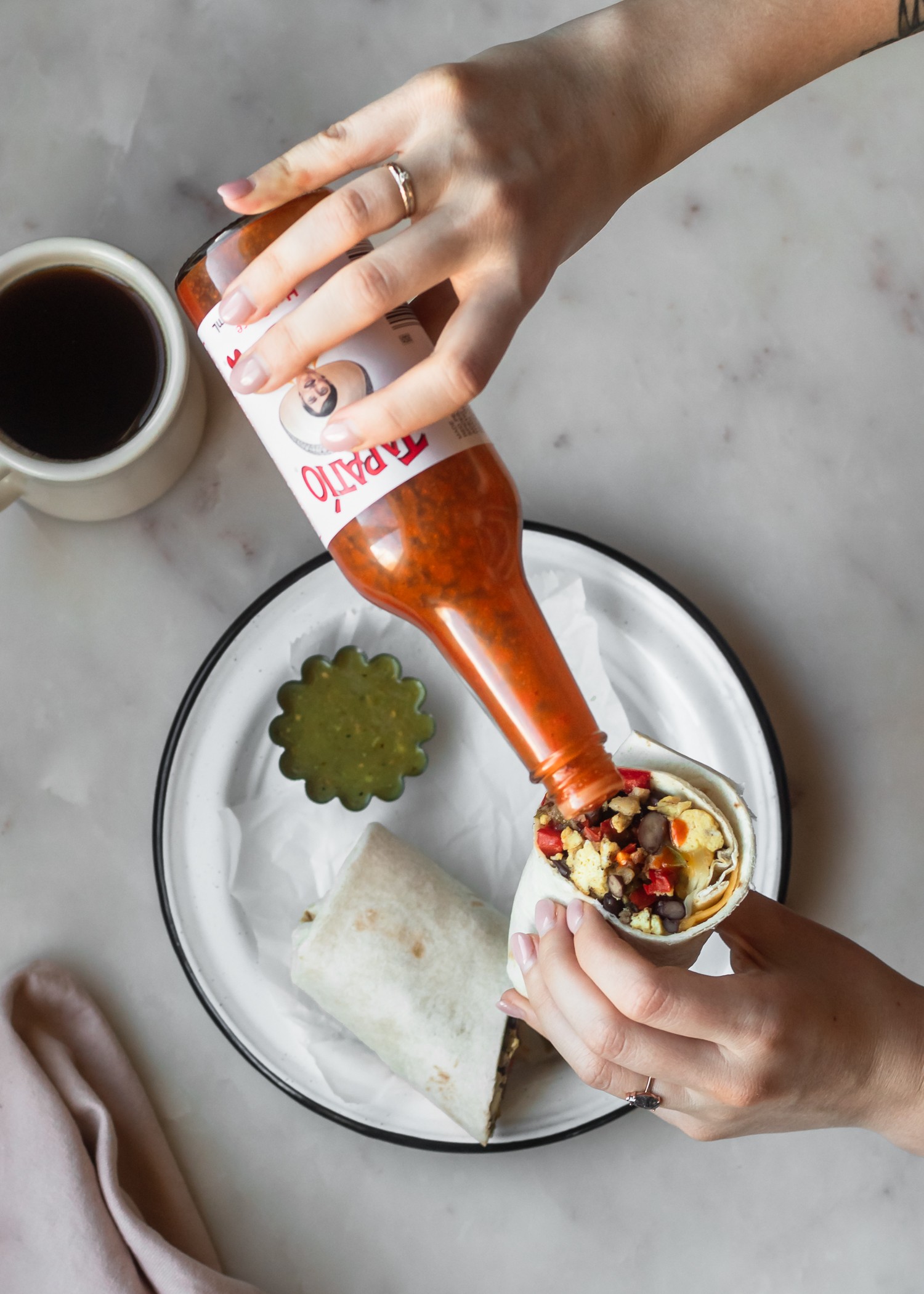 An overhead shot of a woman's hand pouring hot sauce on a breakfast burrito over a white marble counter.