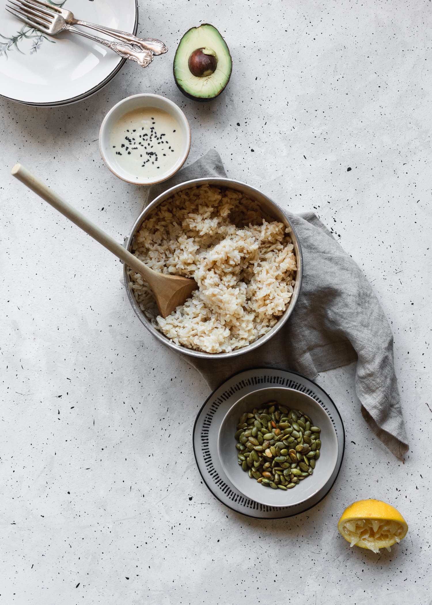An overhead shot of a pot of rice placed on a linen, bowl of pepitas, bowl of tahini dressing, avocado, bowls, and half a lemon in a curved shape on a grey speckled counter.