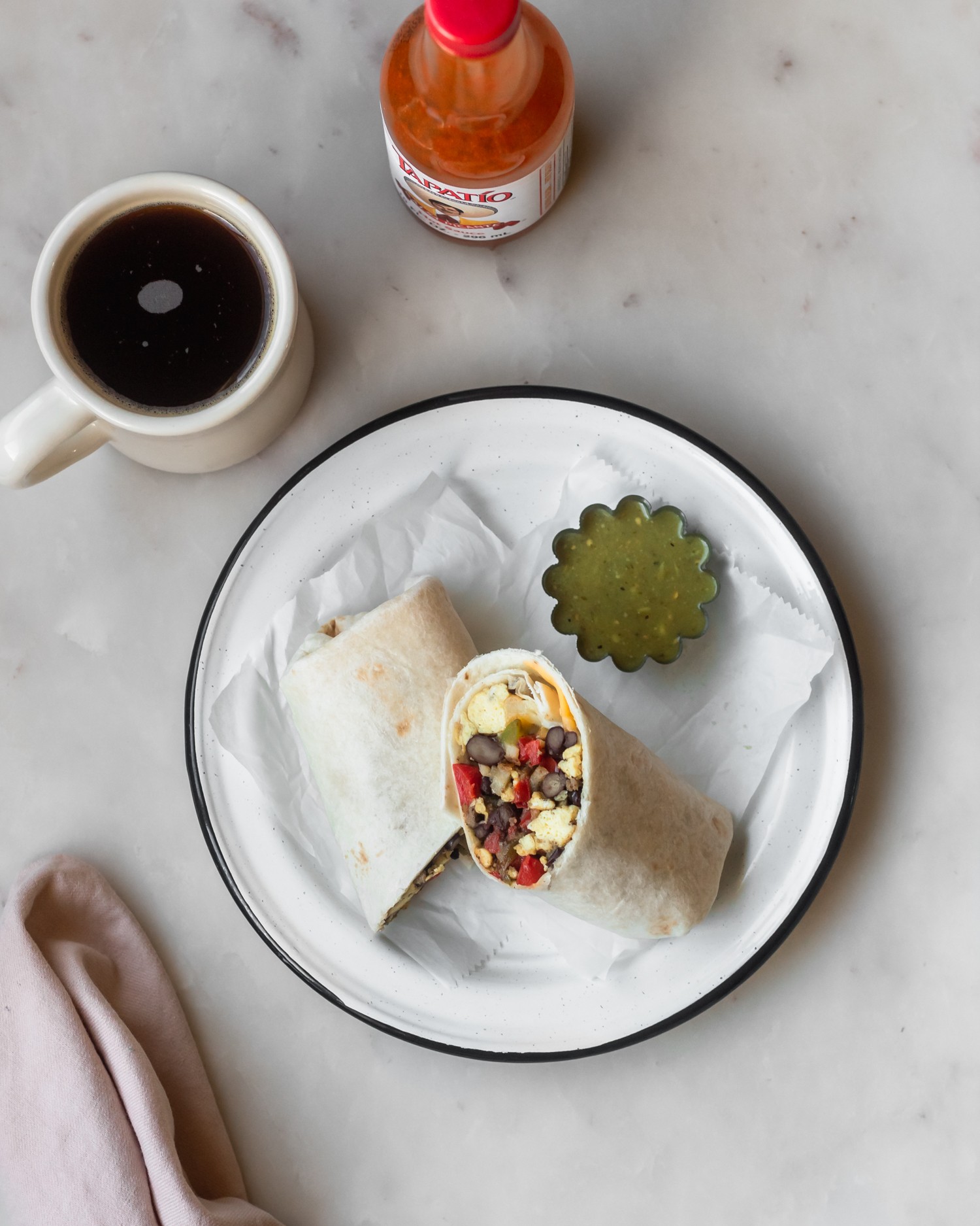 An overhead shot of a breakfast burrito on a white plate with a black rim, with a side of green salsa. The plate is on a white marble counter with a cup of coffee and hot sauce.