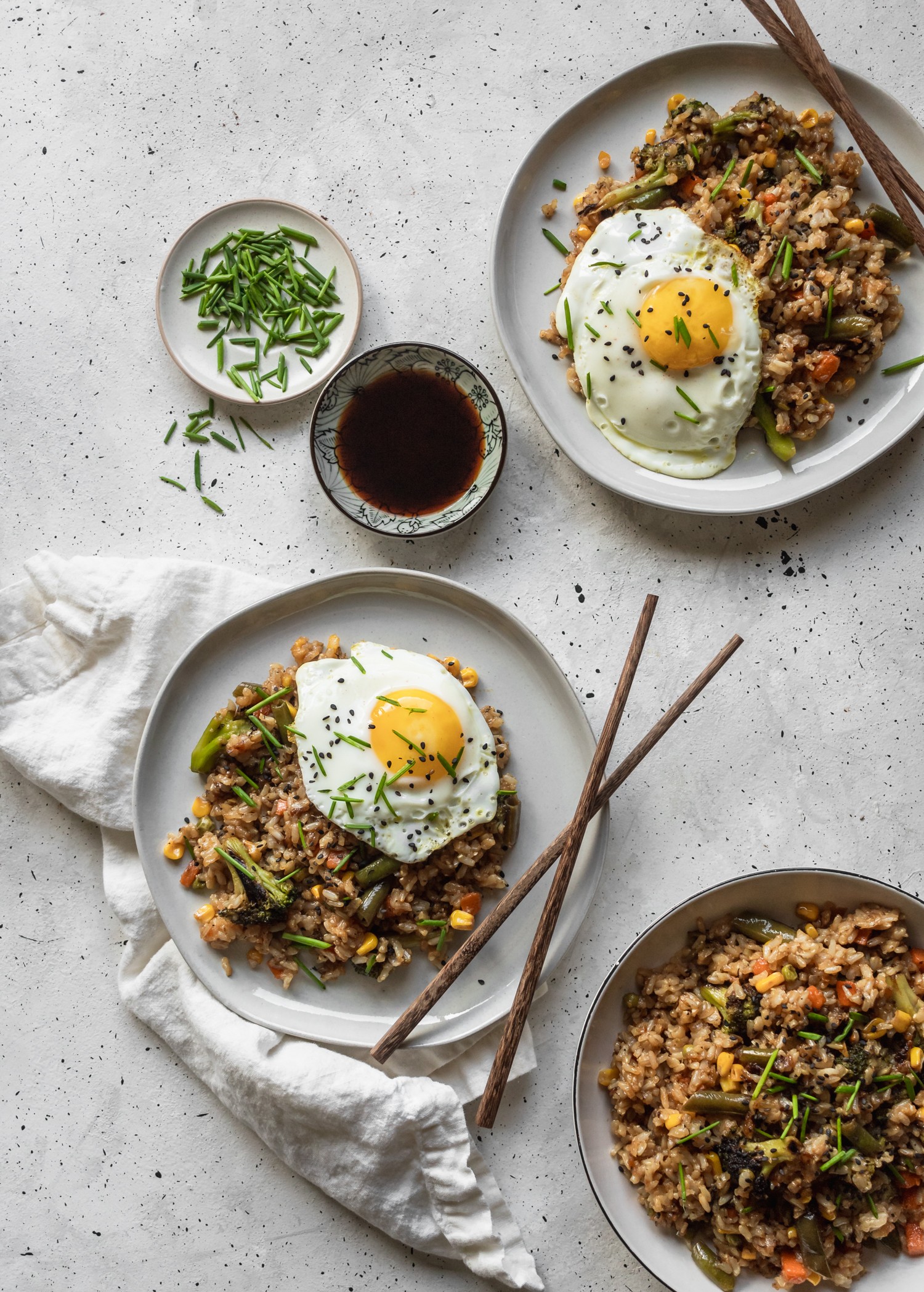 A bird's eye view of three plates of 
 vegetable fried rice with sunny-side up eggs with chopsticks, a white linen. a plate of soy sauce, and a plate of sliced chives on a grey table.