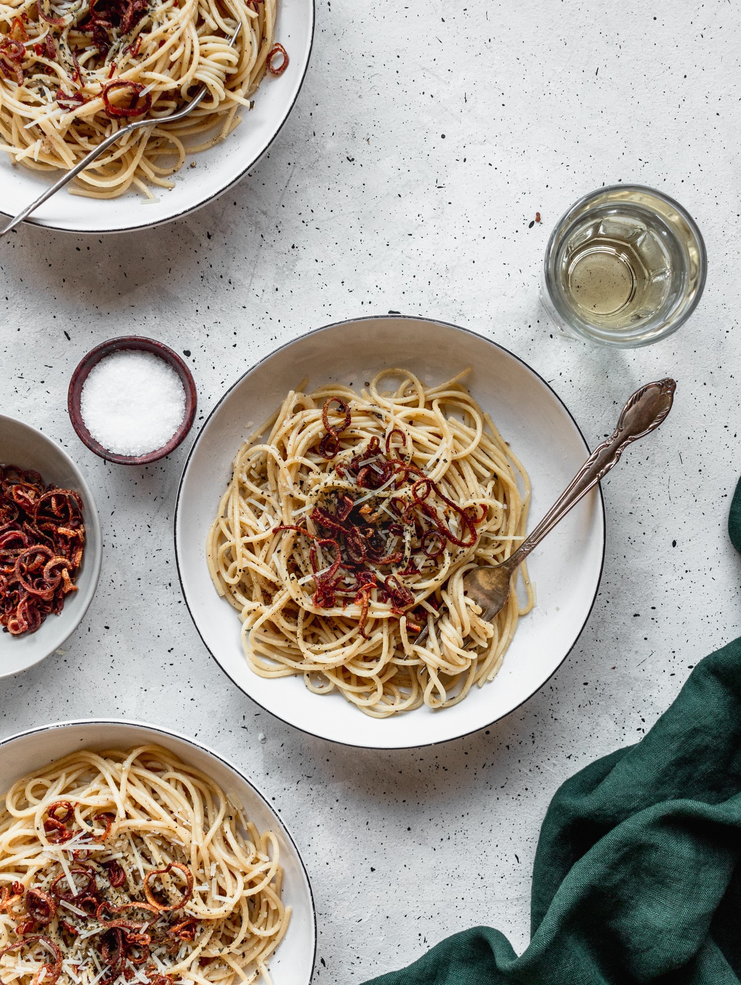 An overhead view of three bowls of pasta on a grey table surrounded by a dark green linen, a glass of white wine, and a bowl of fried shallots.
